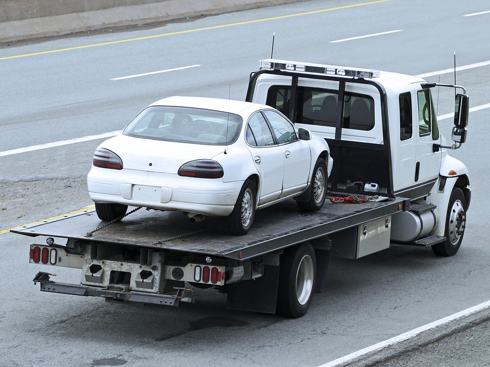 White car on a flatbed tow truck traveling on a gray highway.