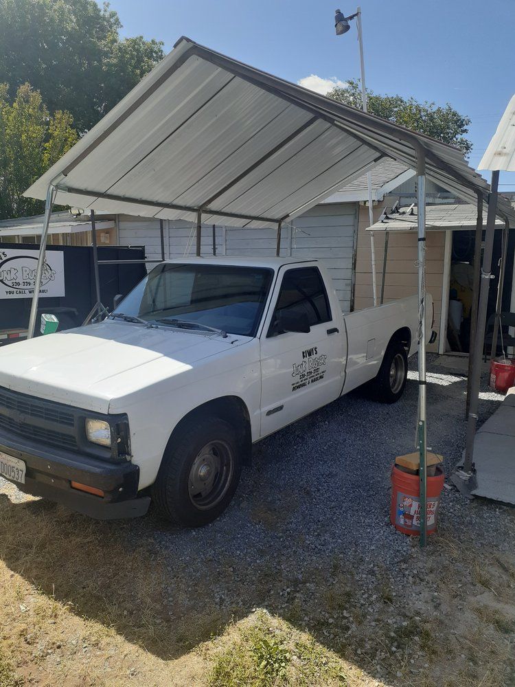 White pickup truck parked under a metal carport in a yard, with a red bucket nearby.