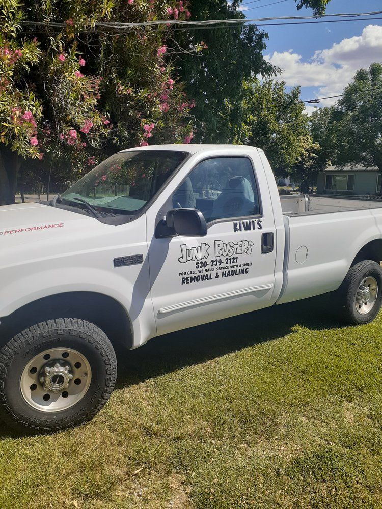 White pickup truck with business logo parked on grass beside flowering trees under a cloudy sky