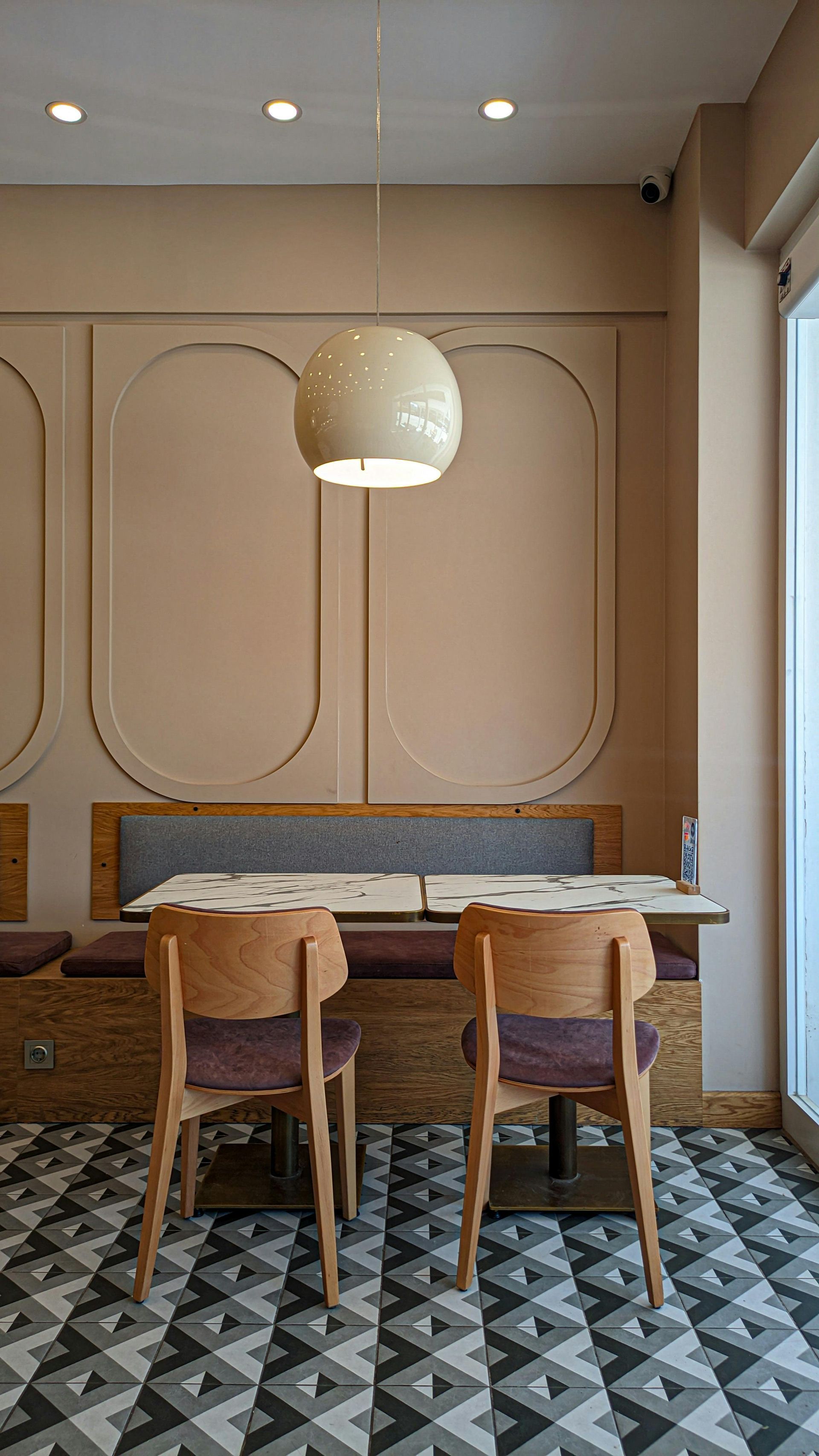 Restaurant seating: two wooden chairs face a table, under a white globe light. Pink wall, patterned floor.