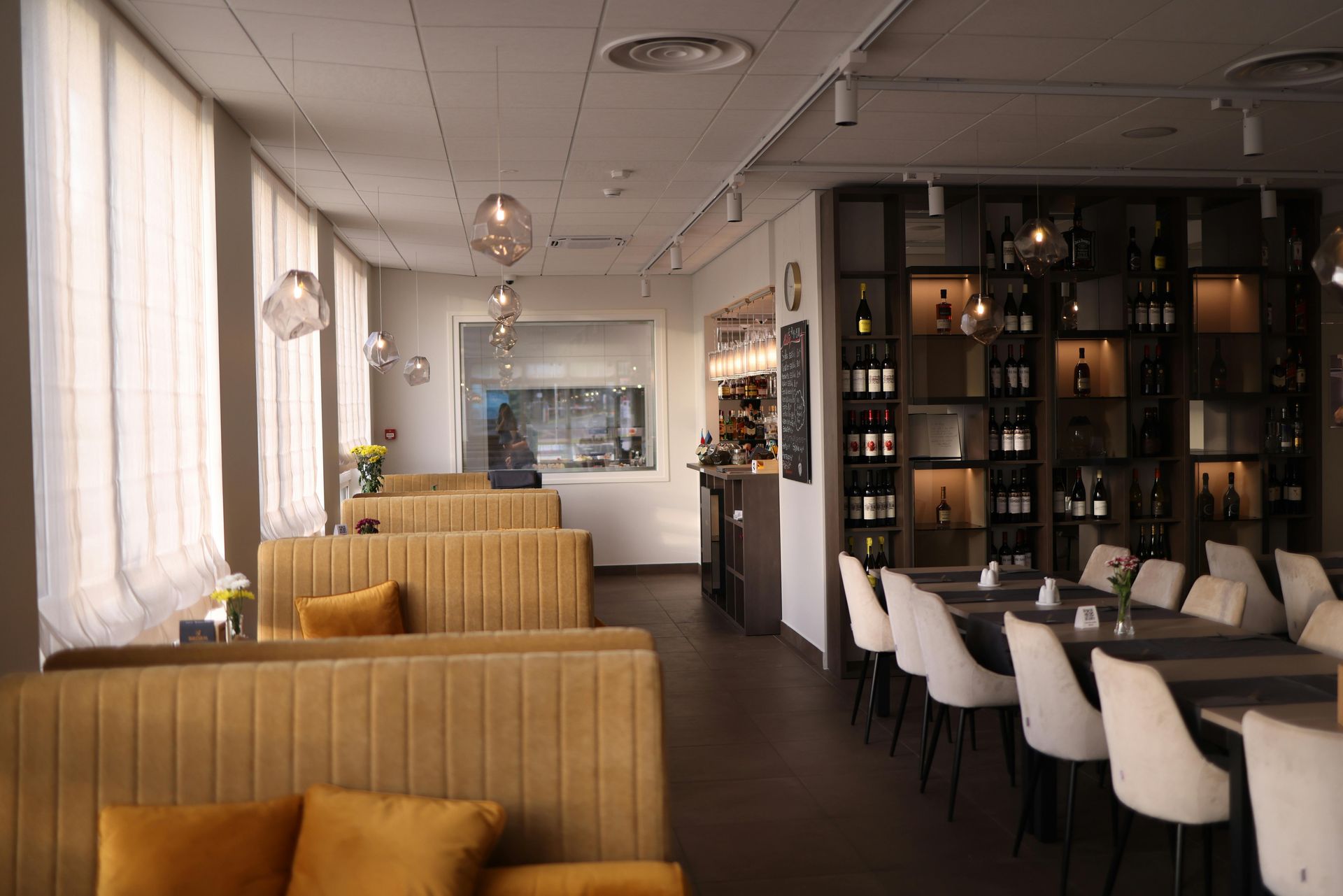 Restaurant interior: mustard-colored booth seating, tables, wine rack, and window with sheer curtains.