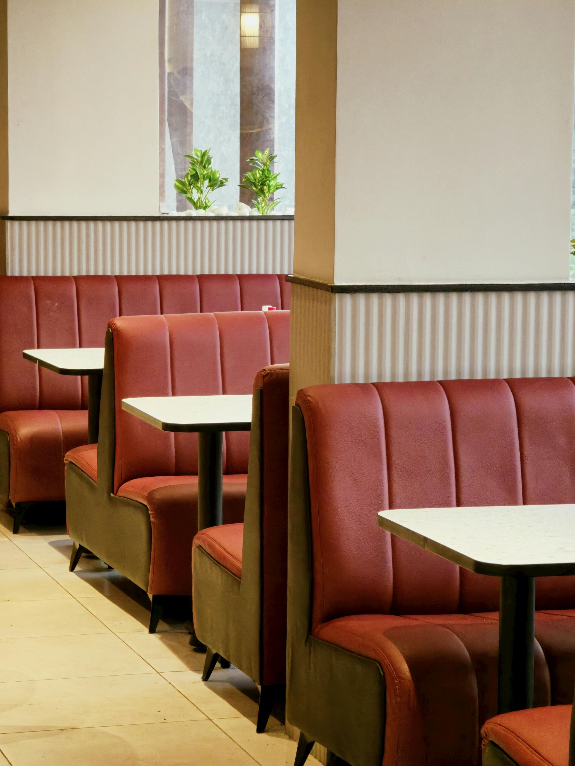 Red booths and tables in a diner, with a white wall and window with plants in the background.