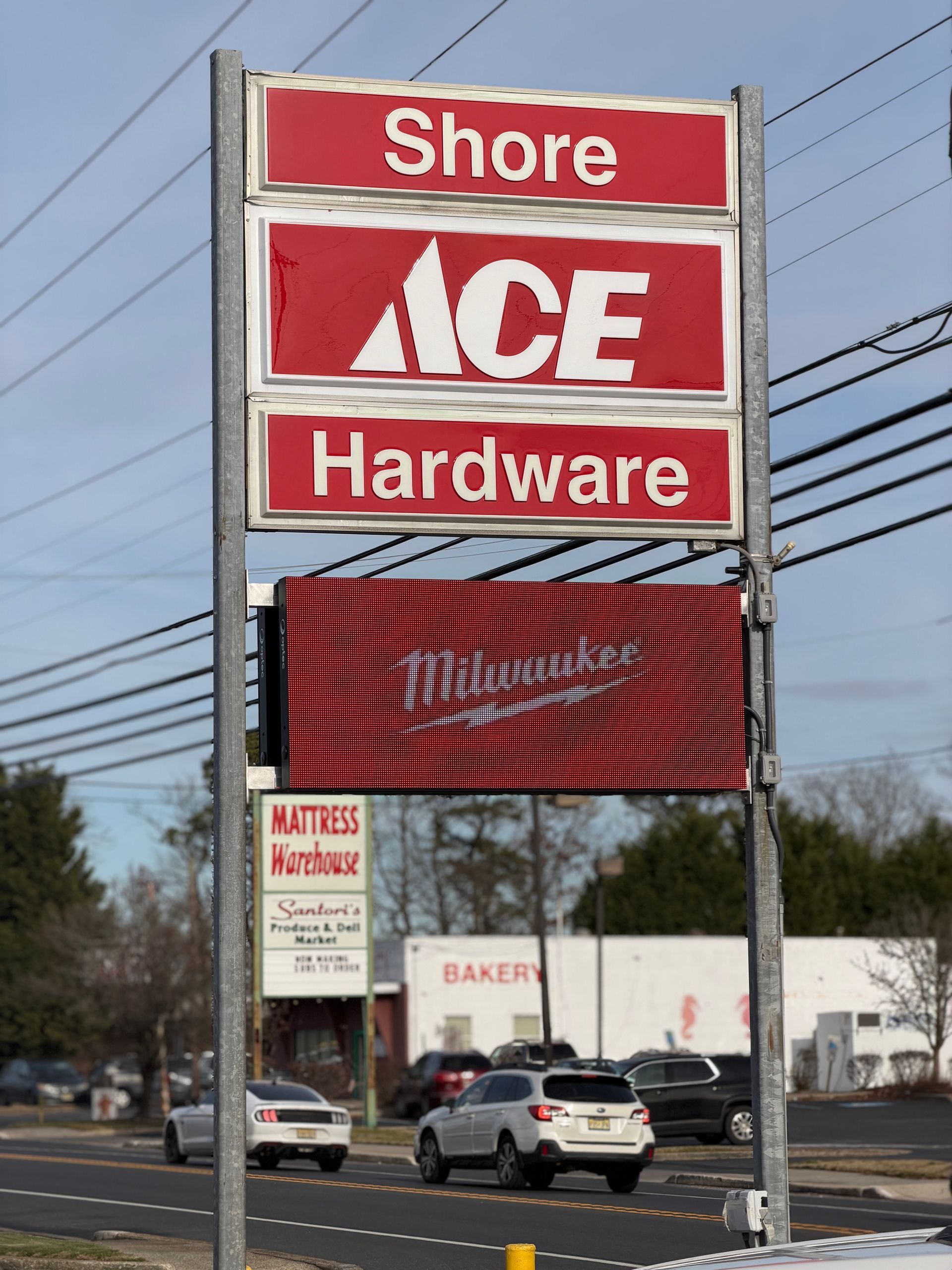 Sign for Shore Ace Hardware with adjacent buildings and passing vehicles.