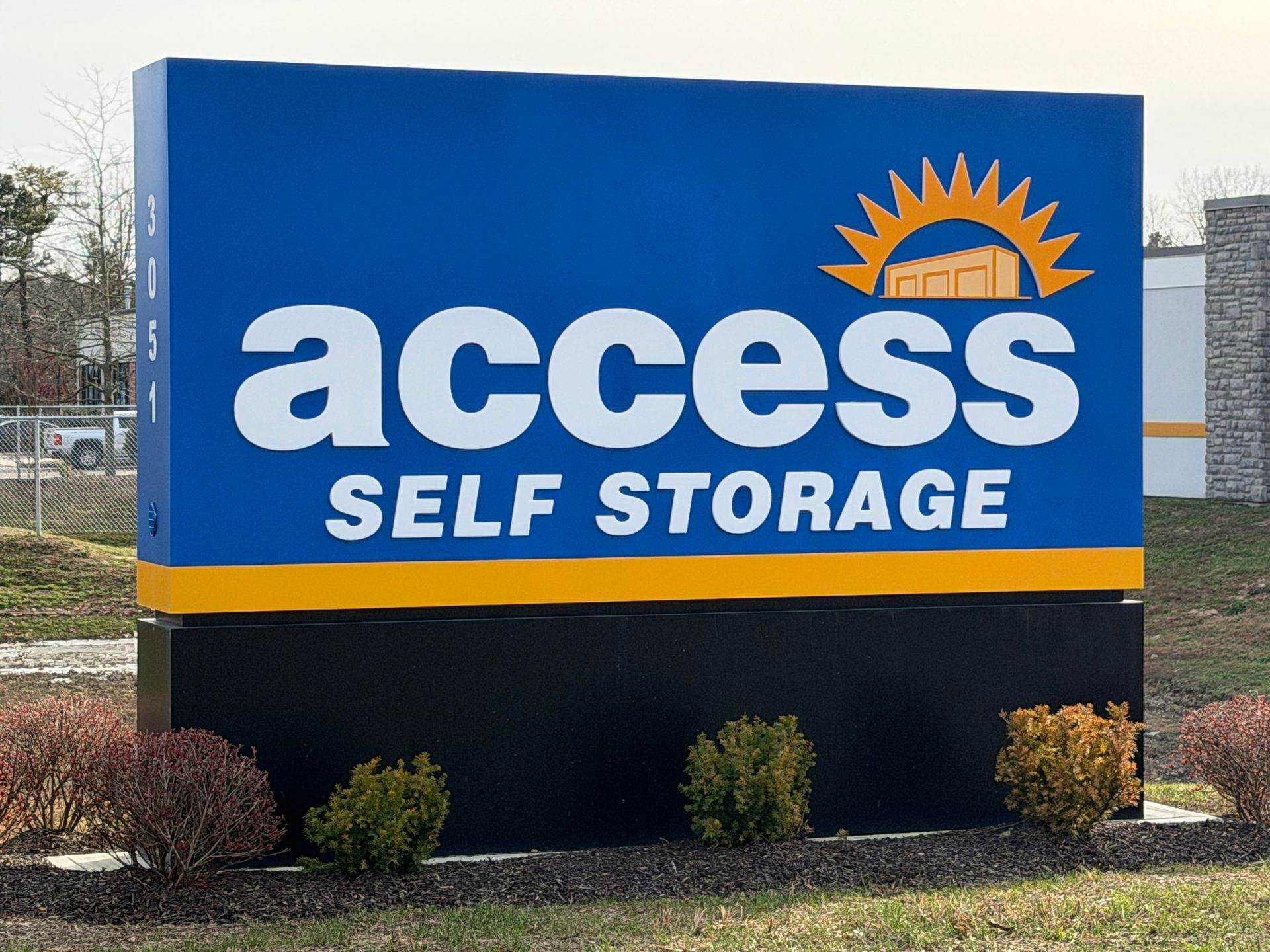 Blue and gold Access Self Storage sign with logo and name, black base, bushes.