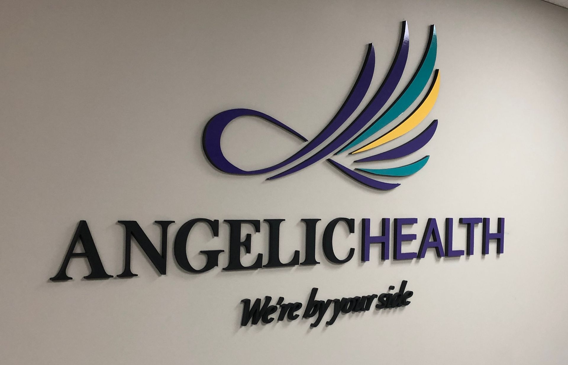 Angelic Health logo on a white wall; features an abstract wing design in blue, teal, and yellow with black text.