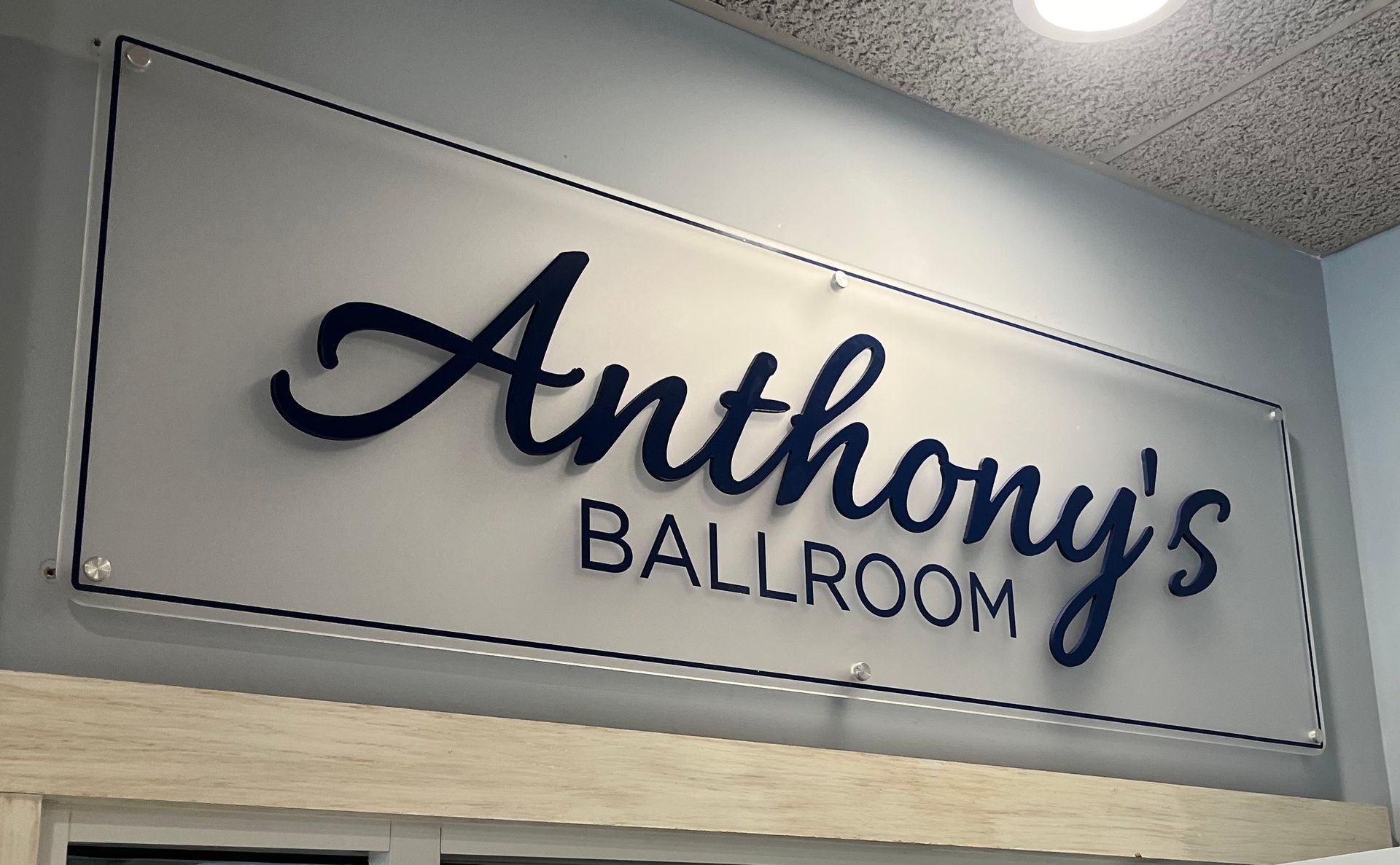 Sign for Anthony's Ballroom, navy blue script lettering on clear background, mounted on a gray wall.
