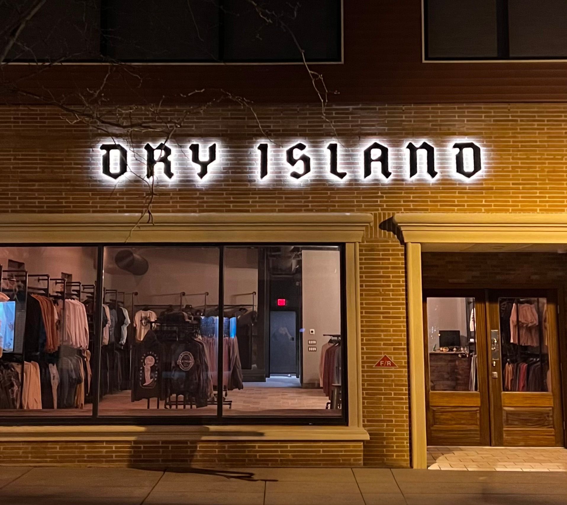 Dry Island storefront with lit-up sign above large windows showcasing clothing displays. Exterior is brick.