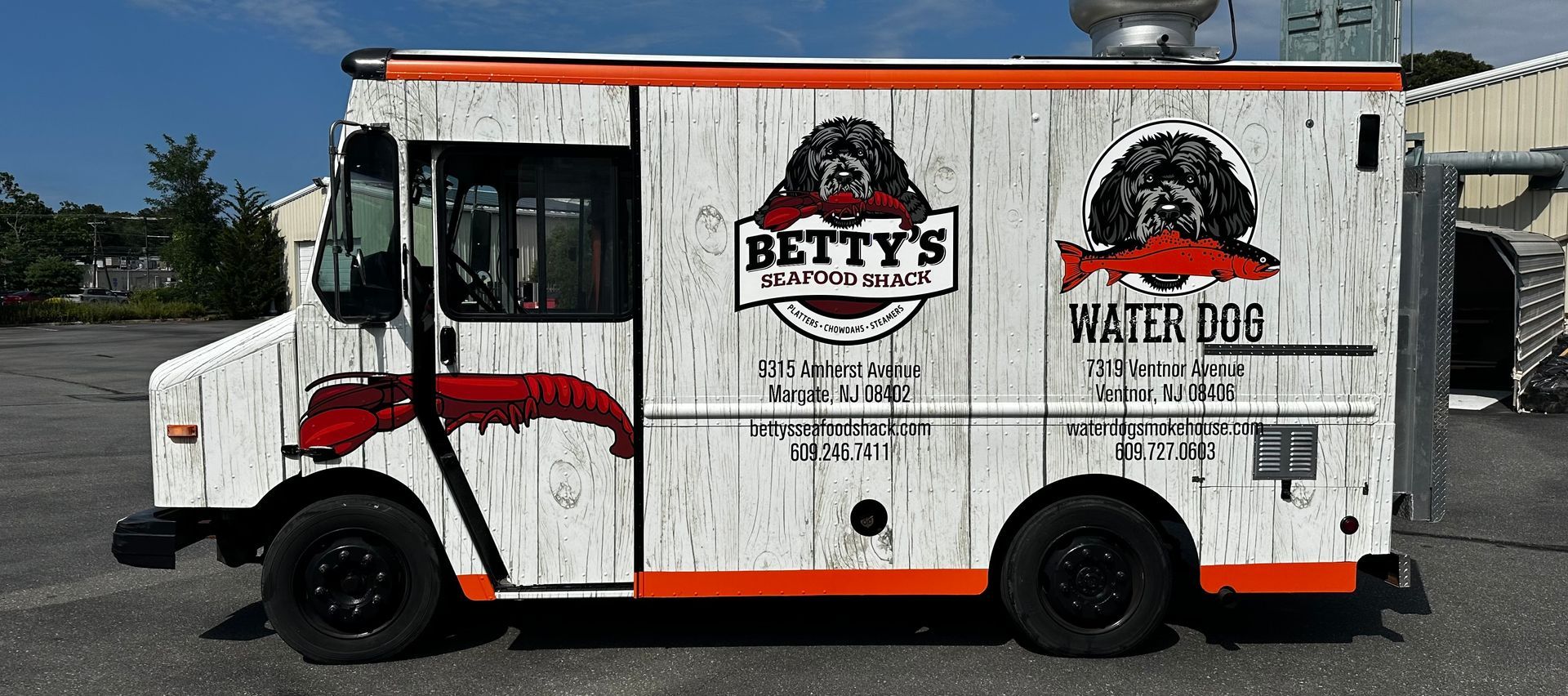 Food truck with white wooden siding, orange trim, and black wheels. It has logos for