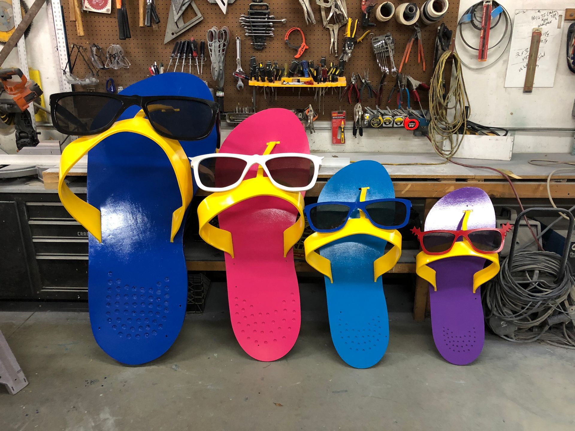 Four colorful flip-flop sculptures wearing sunglasses in a workshop setting.