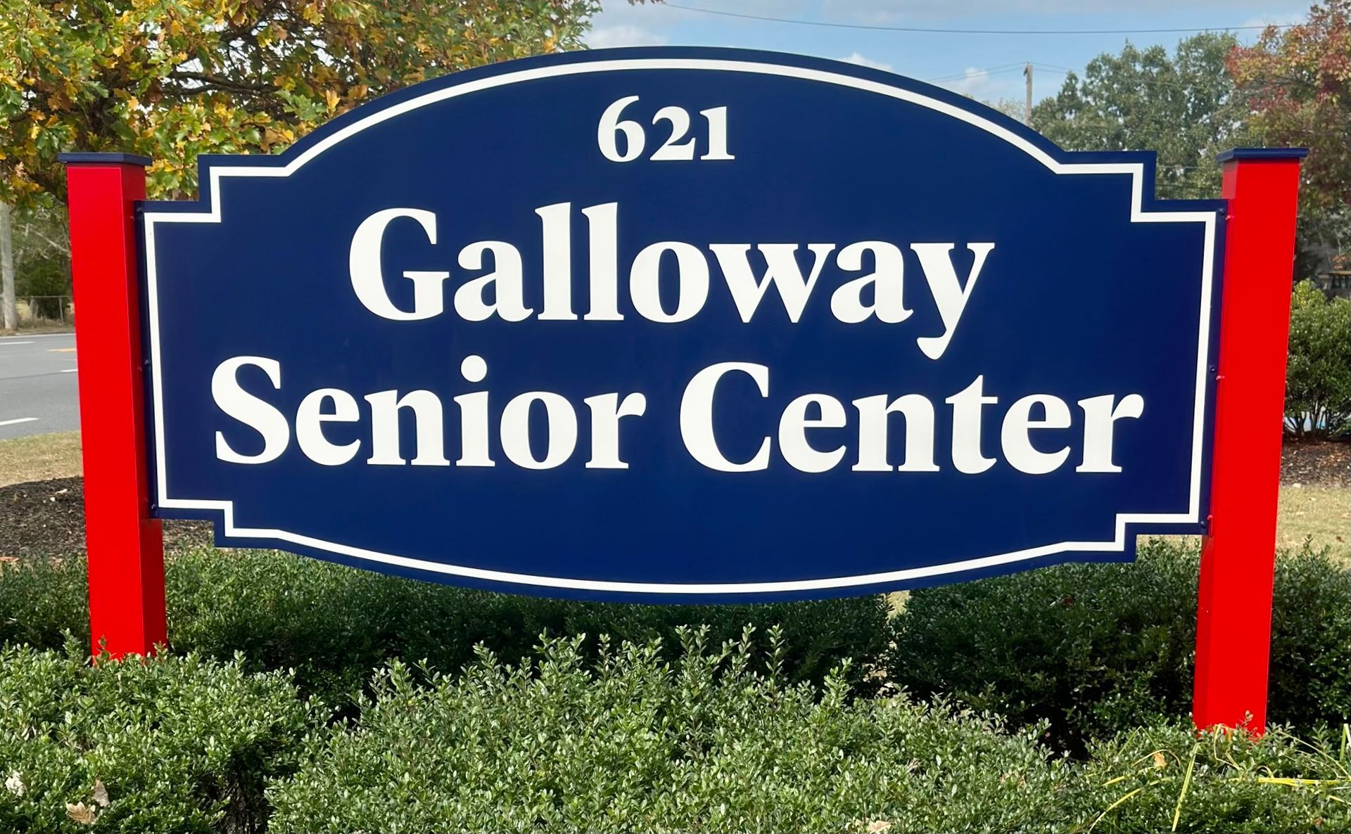 Sign for Galloway Senior Center, blue with white lettering, red posts, bushes.