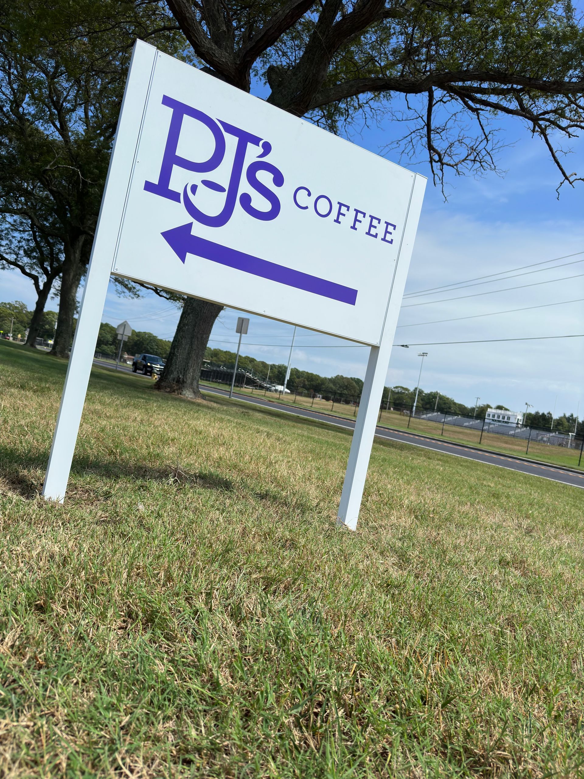 Sign for PJ's Coffee with an arrow pointing left, on a grassy hill with trees and a road in the background.