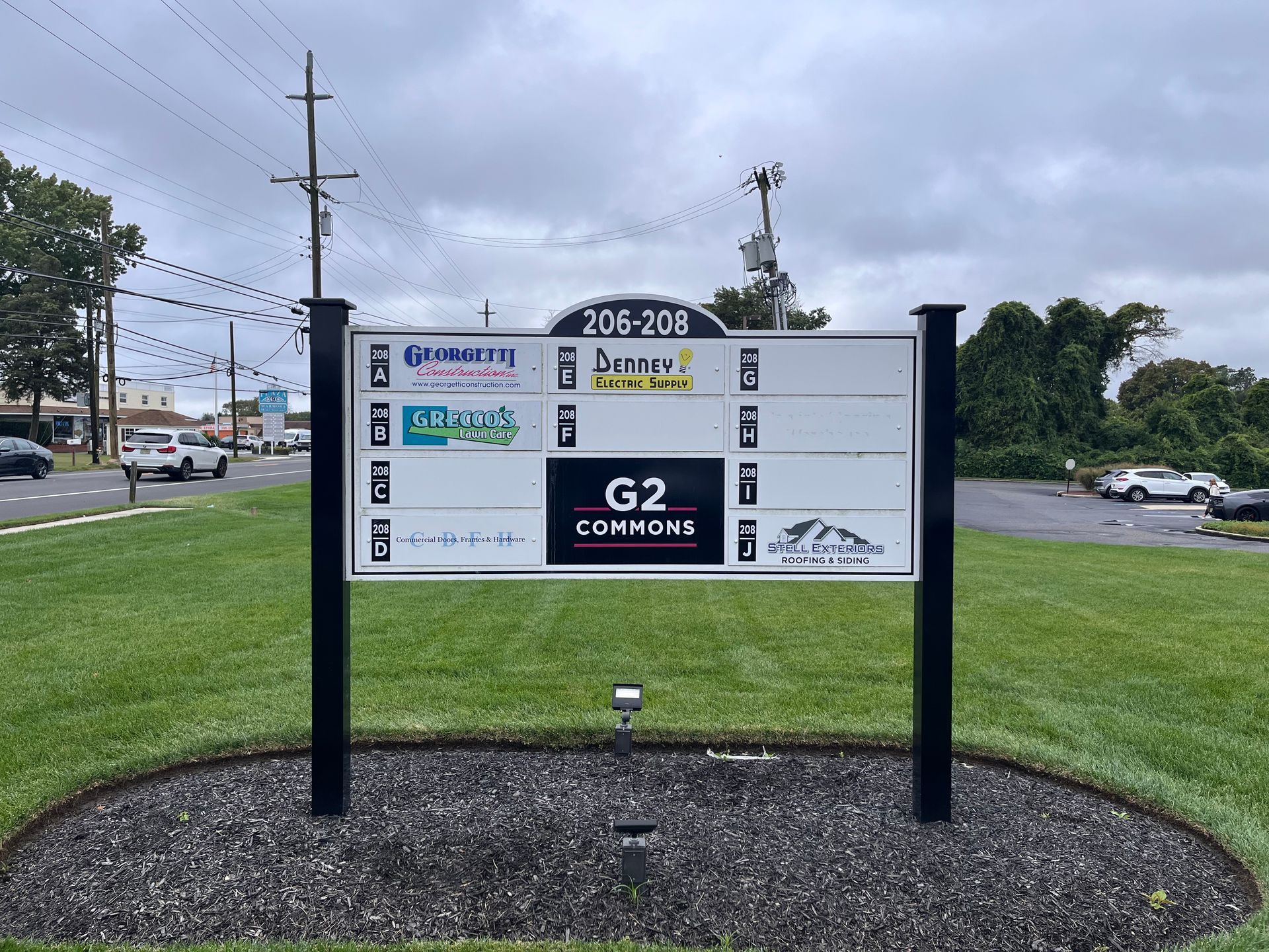Signpost displaying business names at 204-220, black posts on grass, dark mulch base, road and cars visible in background.