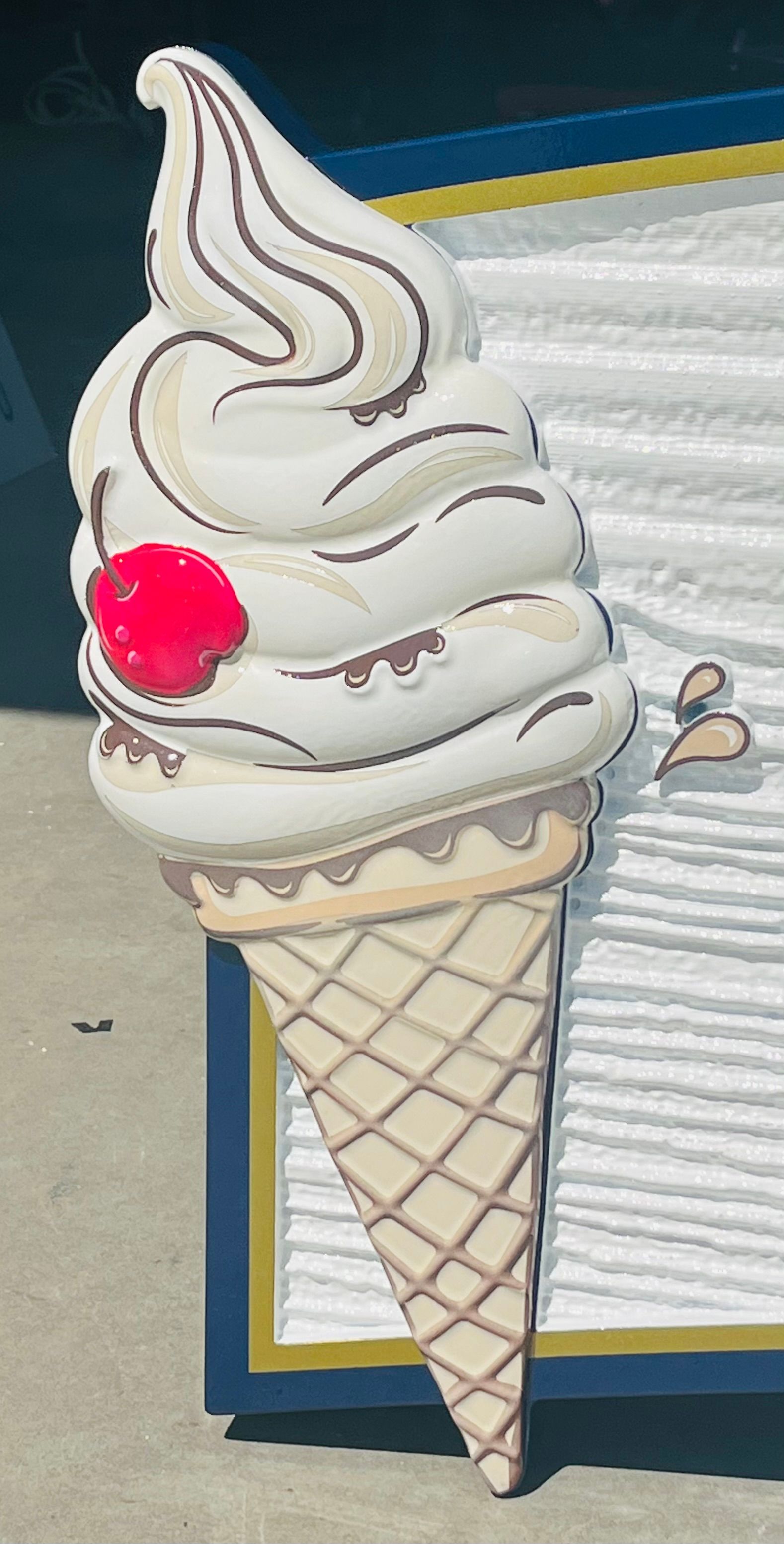 Large, wooden ice cream cone sign with cherry on top; cream, brown, gold, and blue.