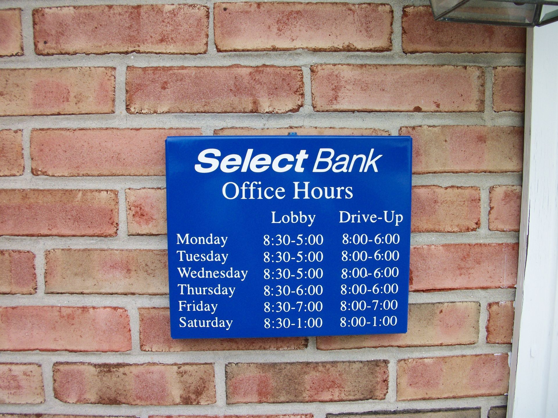 A blue Select Bank office hours sign on a brick wall, listing lobby and drive-up hours for each day.