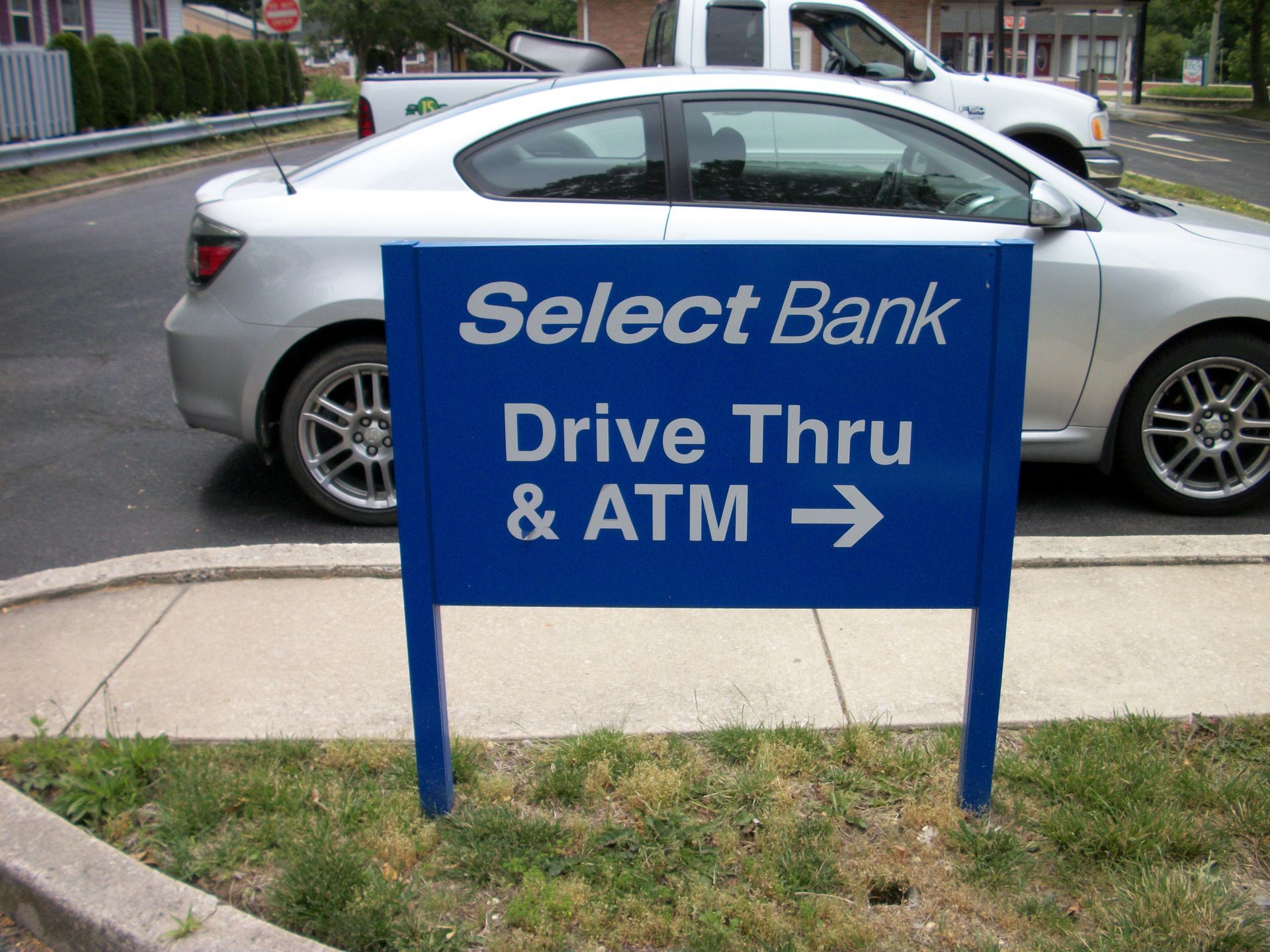 Blue Select Bank drive-thru and ATM sign with a right-pointing arrow. A silver car is parked behind it.