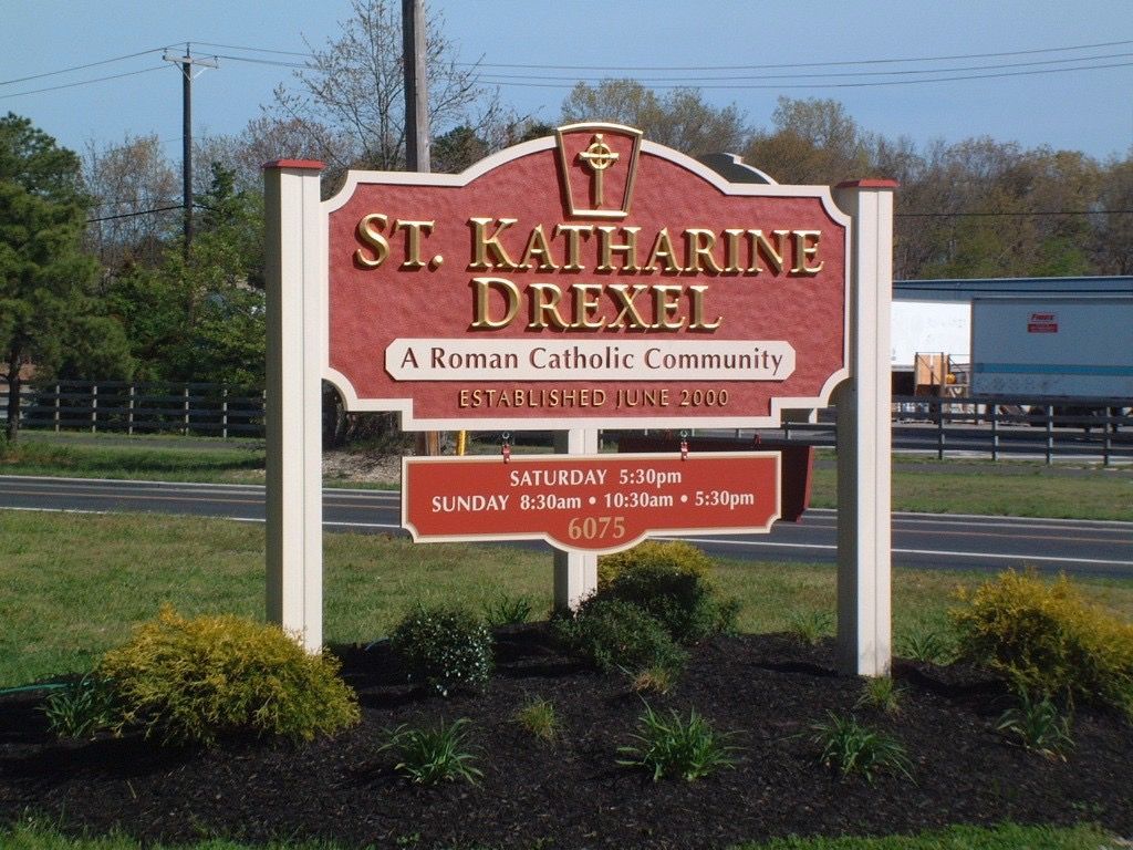 Sign for St. Katharine Drexel Roman Catholic Community, red and cream, with service times visible.