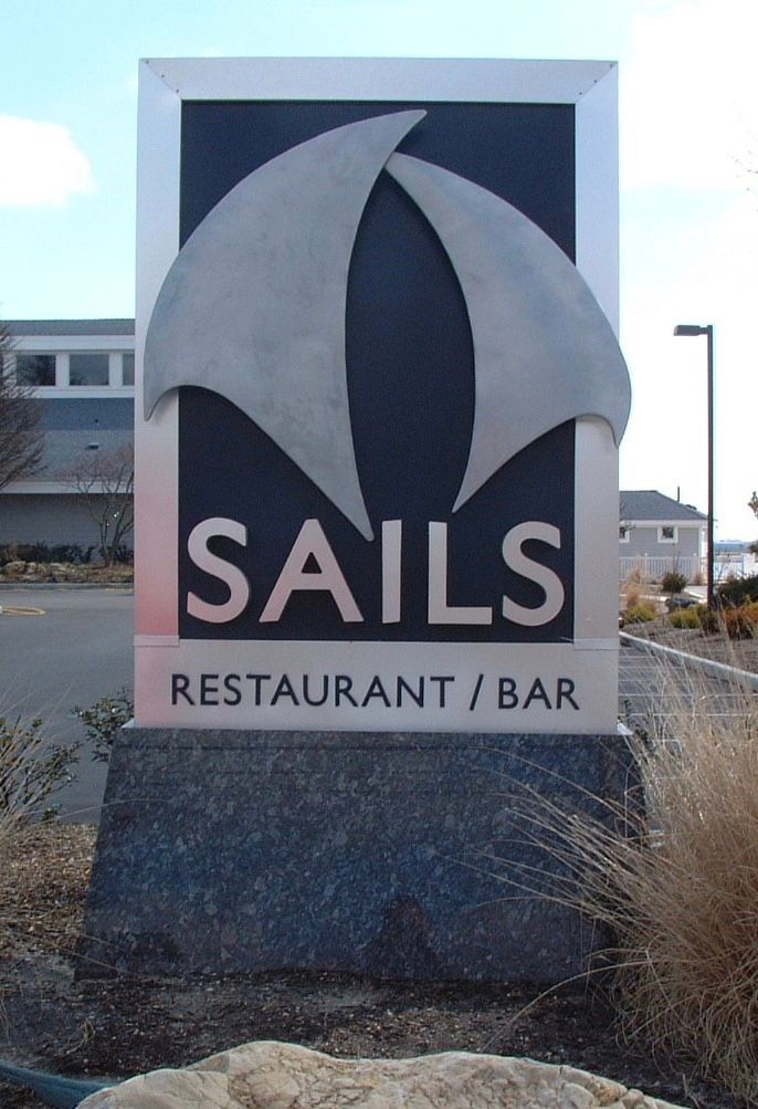 Sign for Sails Restaurant/Bar, with overlapping sail graphic, black background, silver lettering on gray stone base.
