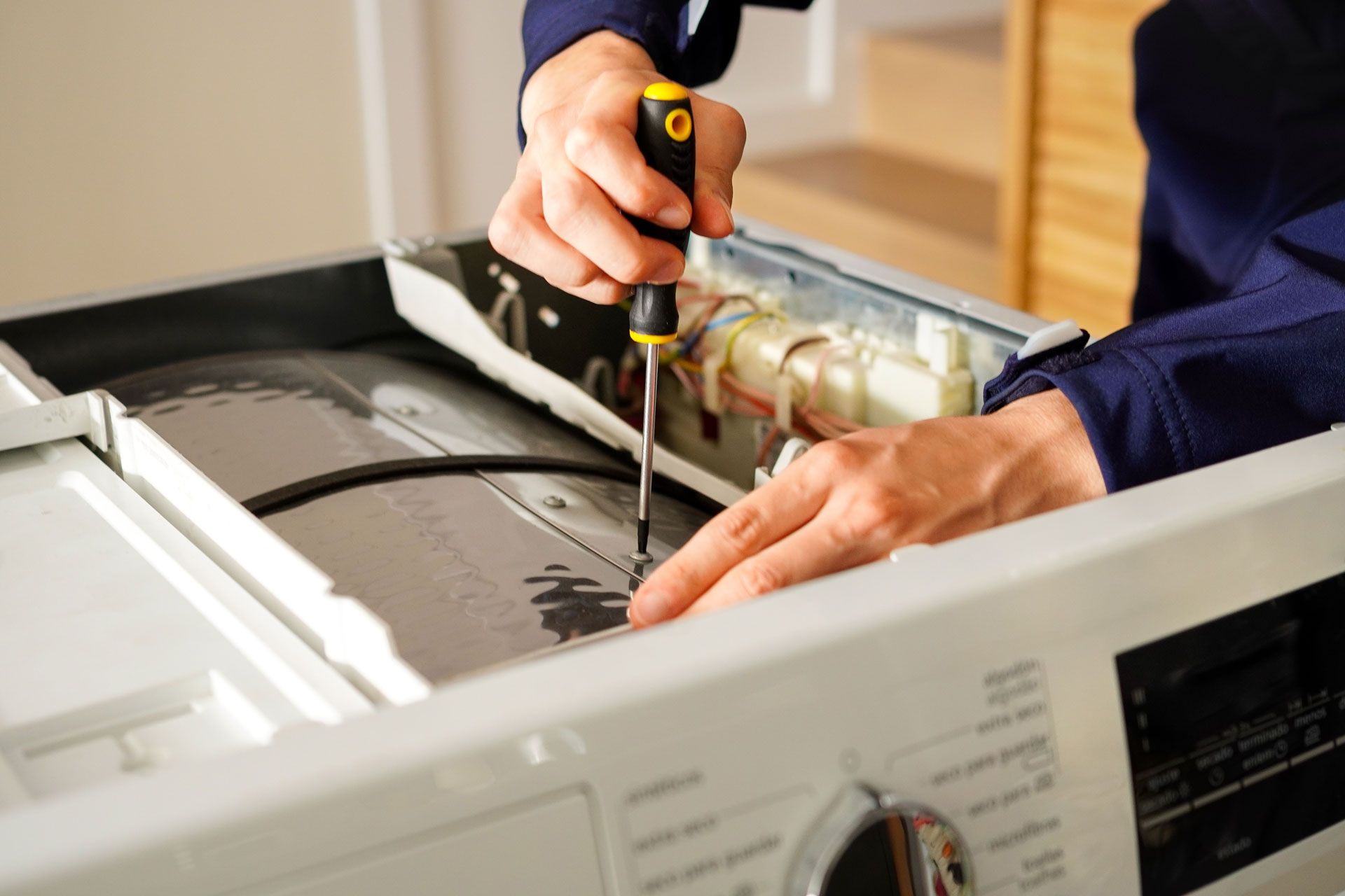 Technician repairing a washing machine with a screwdriver, hands working inside the open appliance.