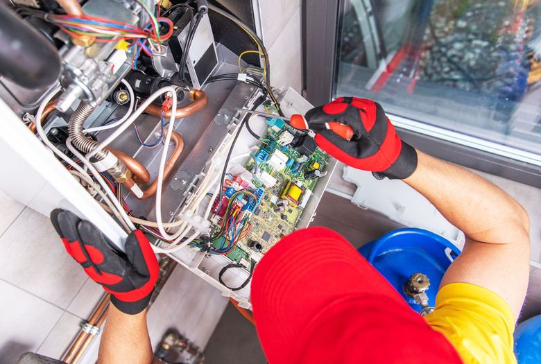 Technician in red shirt and gloves repairing wiring inside an electrical cabinet next to a blue bucket