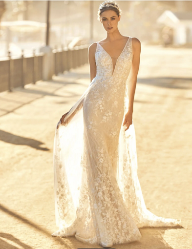 Woman in a wedding dress with a flowing train and sheer cape, standing outdoors in a sunny setting.
