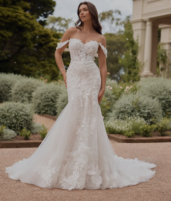 Woman in a white off-the-shoulder wedding dress poses outdoors in a garden setting.