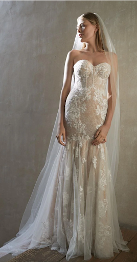 Woman in a white mermaid wedding dress with a long, detailed train. She has hands on hips.