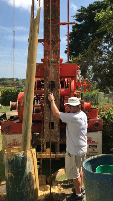Workers inspecting a deep well, using long rods. One wears a blue hard hat and orange shirt.