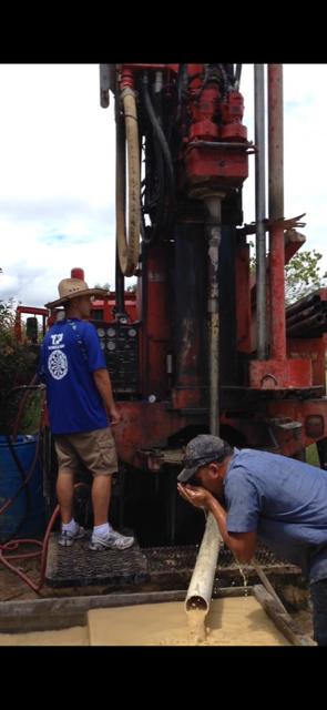 Men in blue shirts operate a drilling rig in a grassy field, trees in background, sunlight.