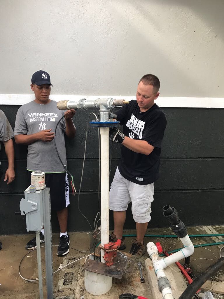 Blue water tank with attached pipes and fittings, next to a black tank, outdoors in a yard.