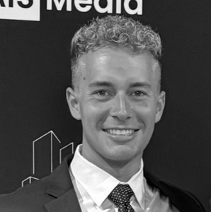 Smiling man in suit, short curly hair, against a dark background with a logo.