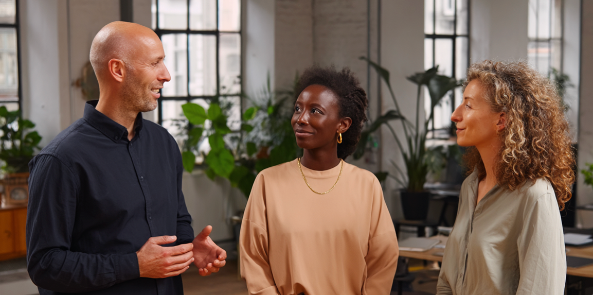 Three diverse colleagues in a bright office conversing. The bald man gestures while the women listen attentively.