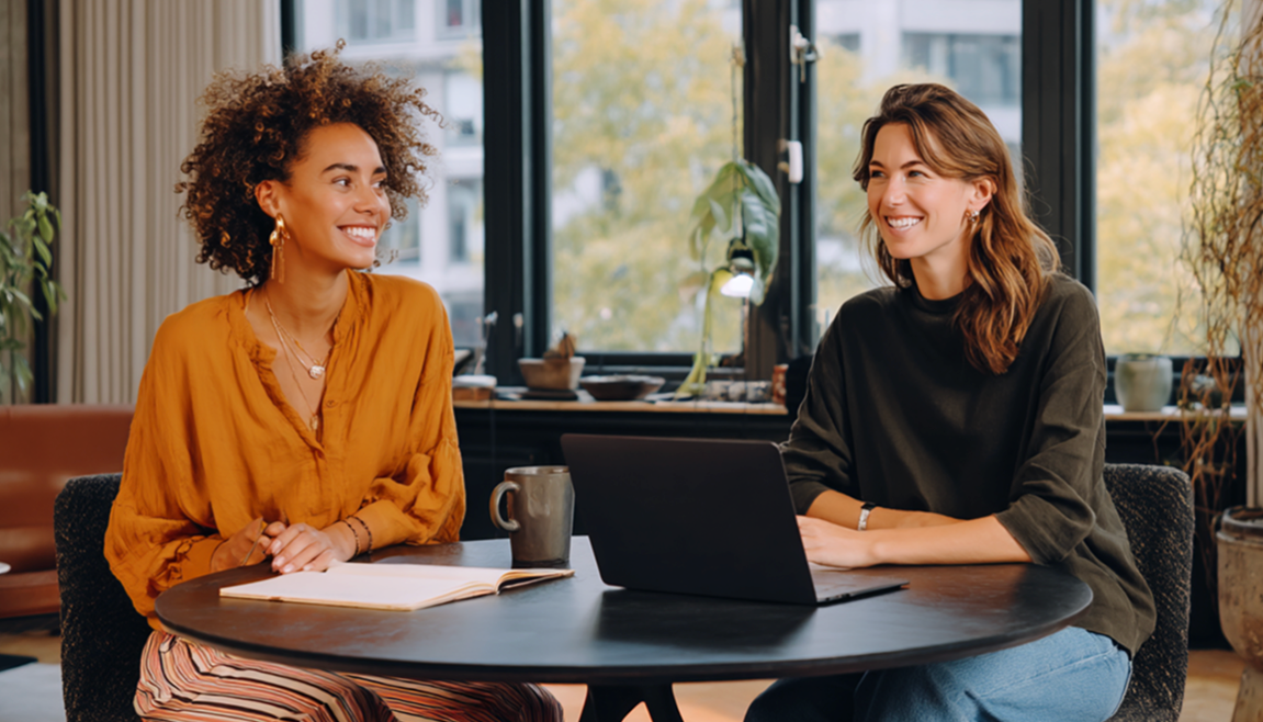 Two women at a round table, smiling and working. One is wearing an orange shirt, the other a green sweater, indoors.