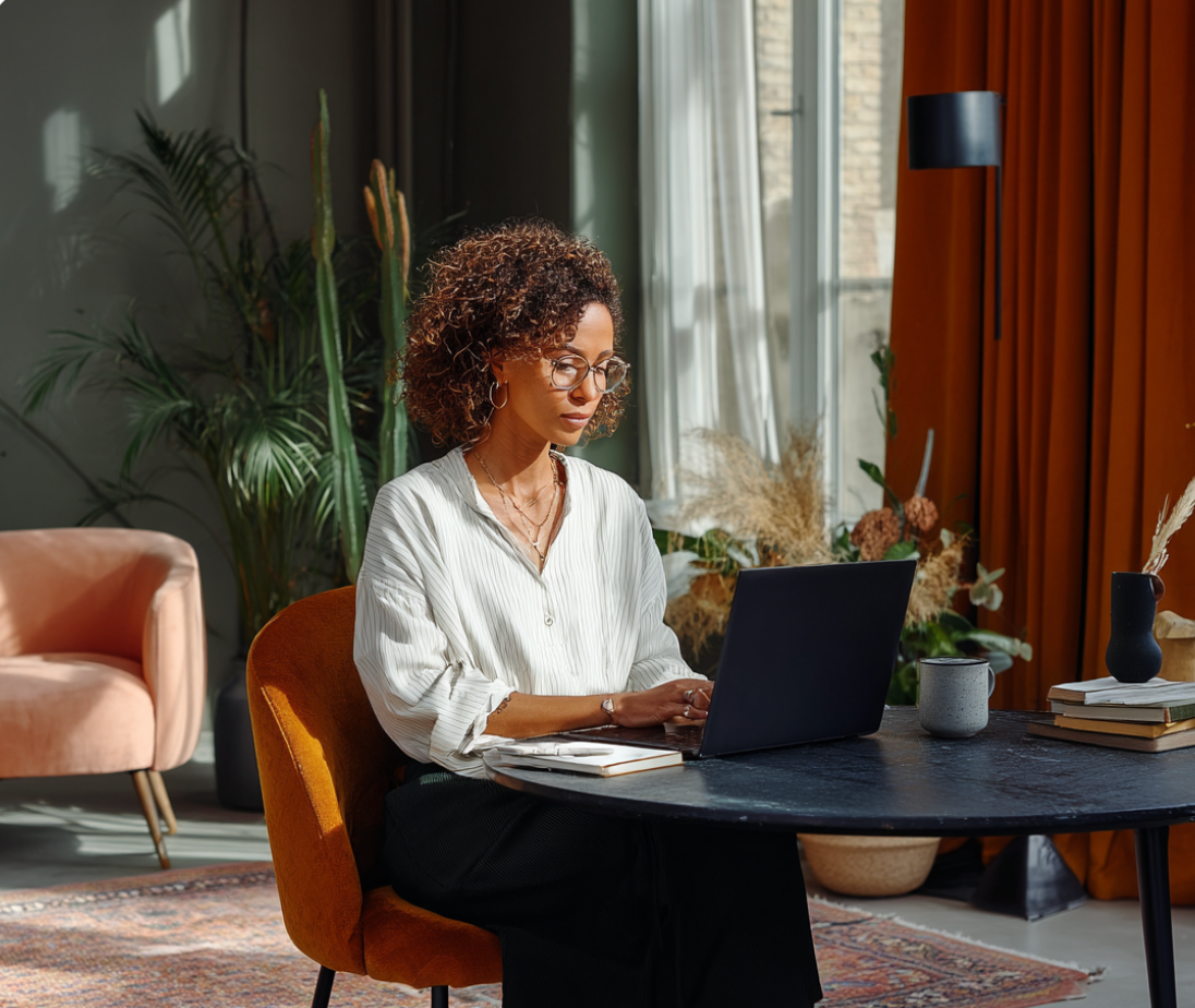 Woman with glasses typing on a laptop at a table, seated in a stylish room with plants and warm lighting.