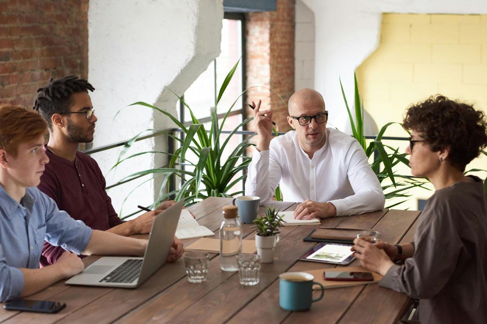 Four people at a table discussing, man gesturing. Laptop, water bottles, and plants present.
