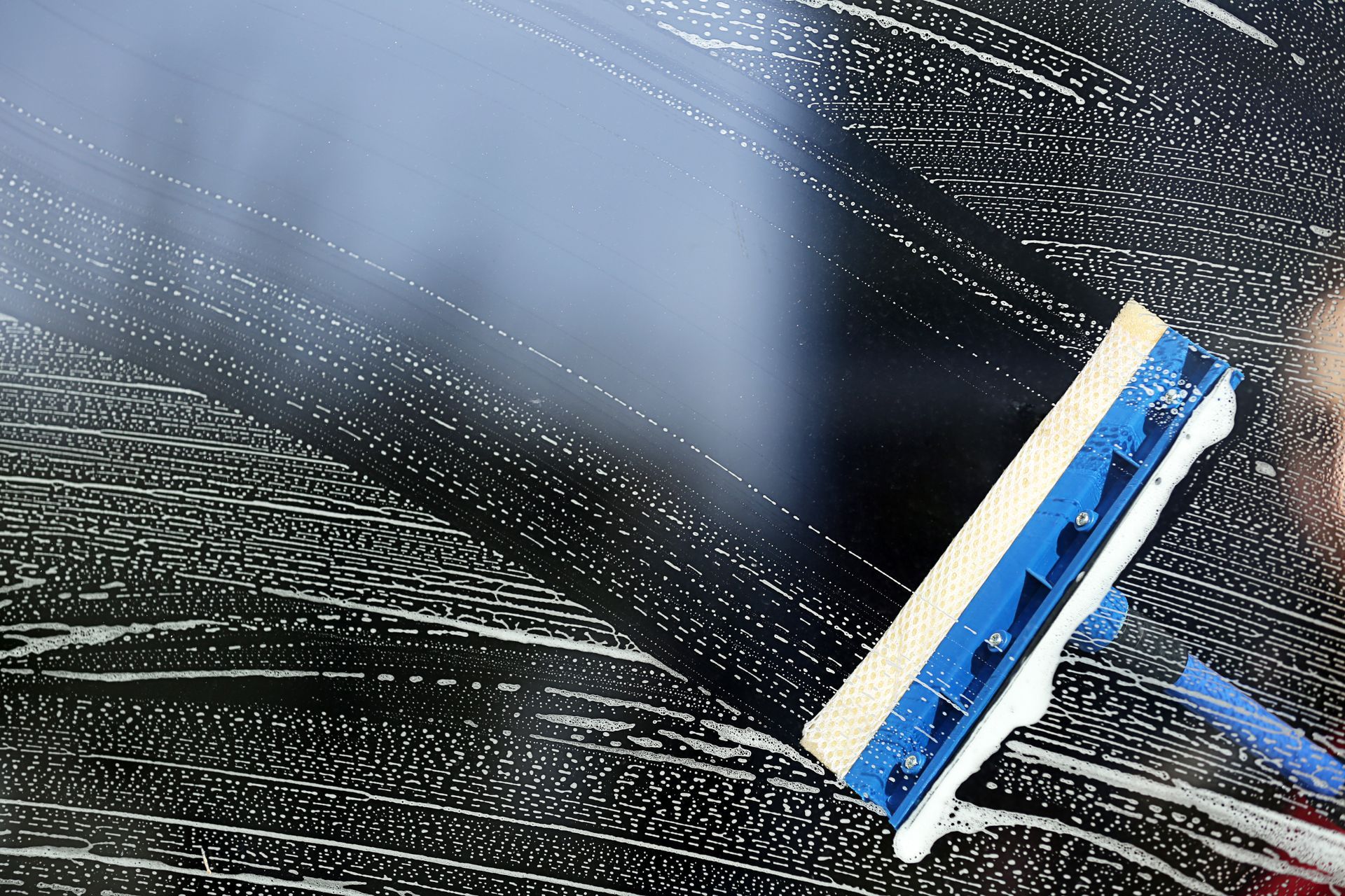 A person uses a blue squeegee with a sponge head to clean soapy foam off a glass surface.