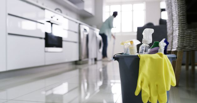 A cleaning bucket with yellow rubber gloves sits in the foreground, with a person cleaning in a blurred kitchen background.