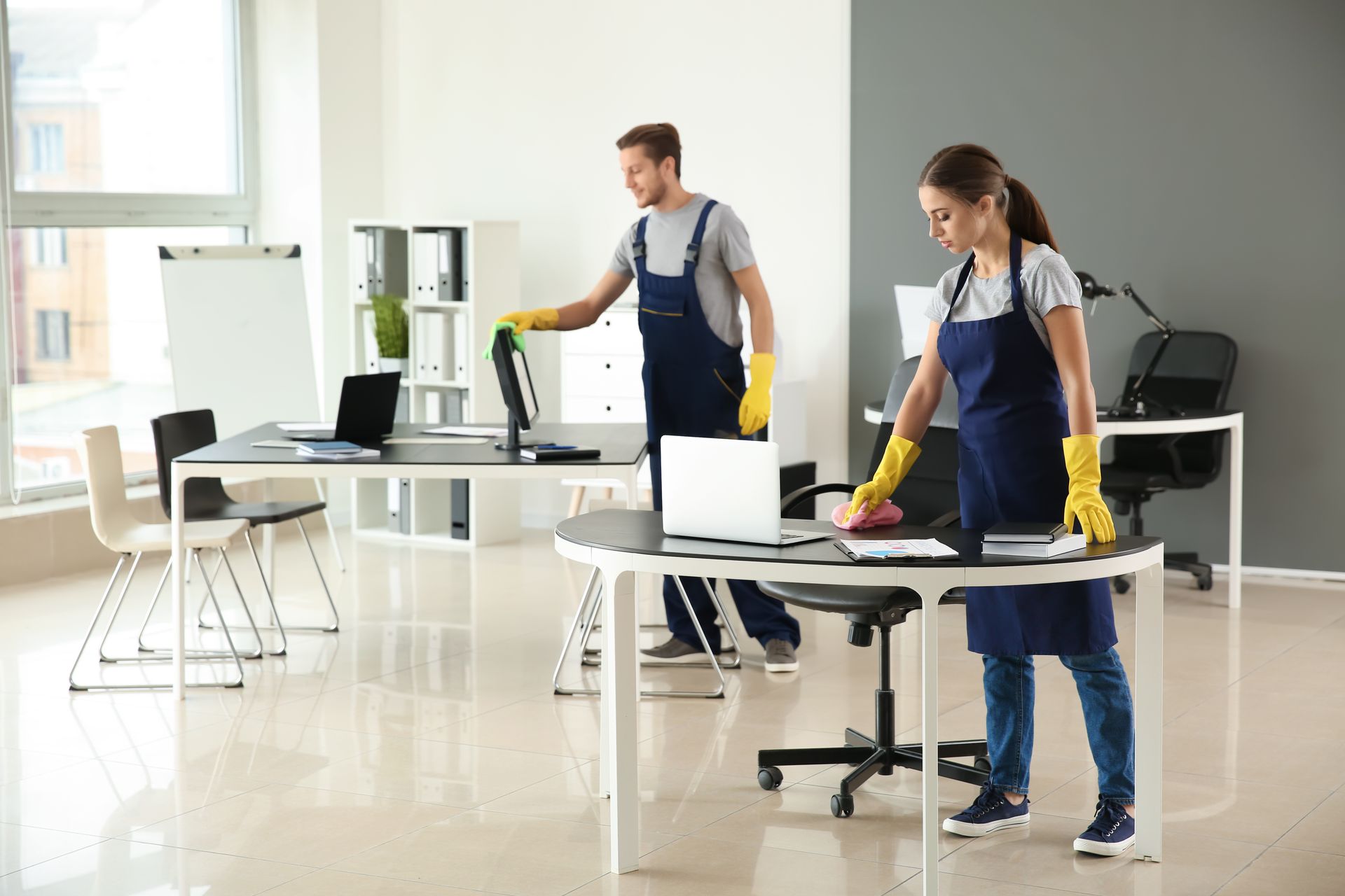 Two professional cleaners in uniforms and yellow gloves clean computer desks in a bright, modern office.