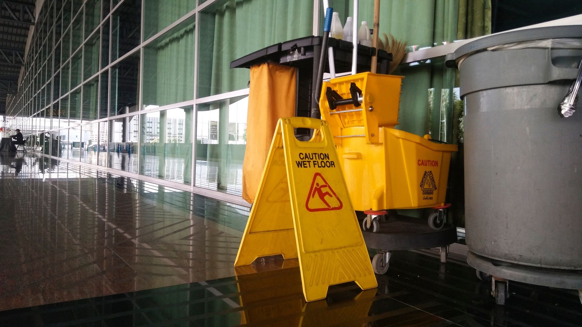 A yellow caution sign stands on a shiny hallway floor near a mop bucket and a gray trash bin.