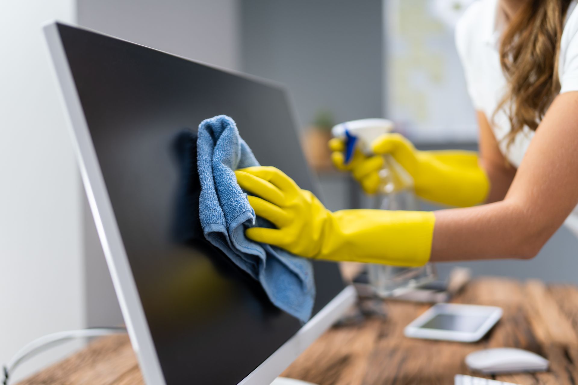 Person wearing yellow rubber gloves cleaning a computer monitor with a blue cloth at a wooden desk.