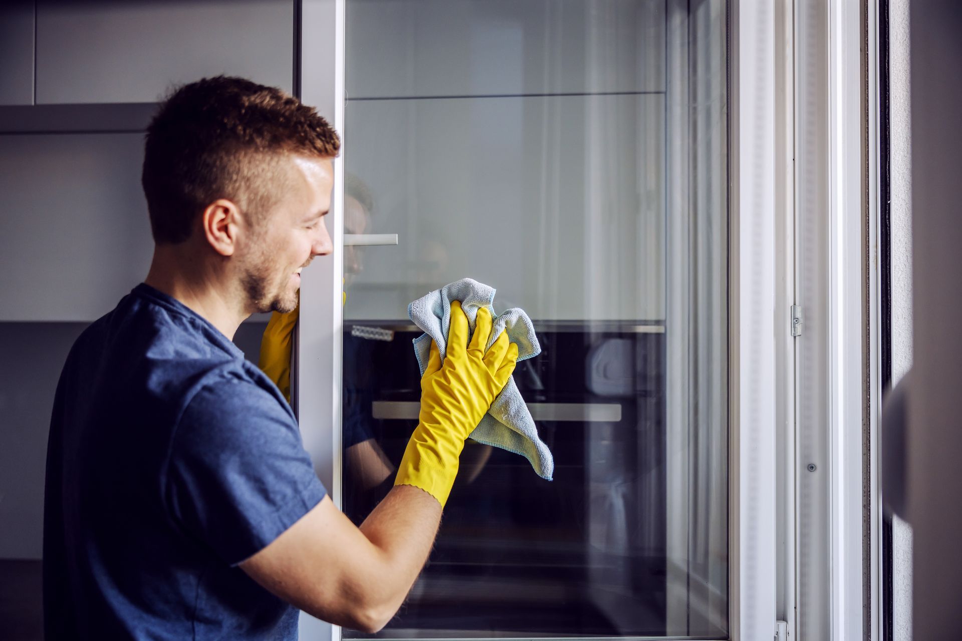 A person wearing a blue shirt and yellow cleaning glove wipes a glass surface with a cloth in a kitchen.