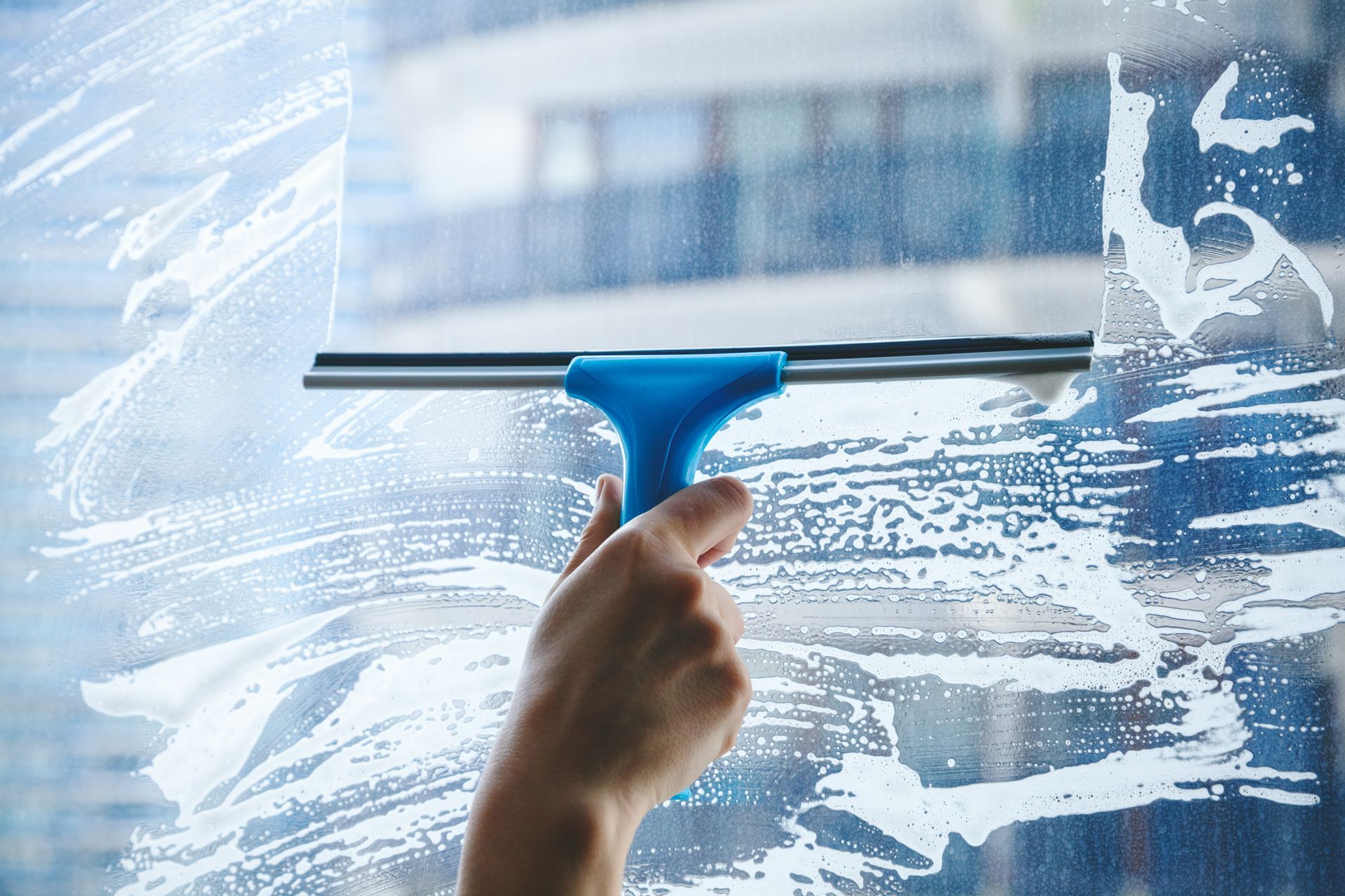 A person uses a blue-handled squeegee to wipe soapy foam off a glass window, revealing a blurry building in the background.