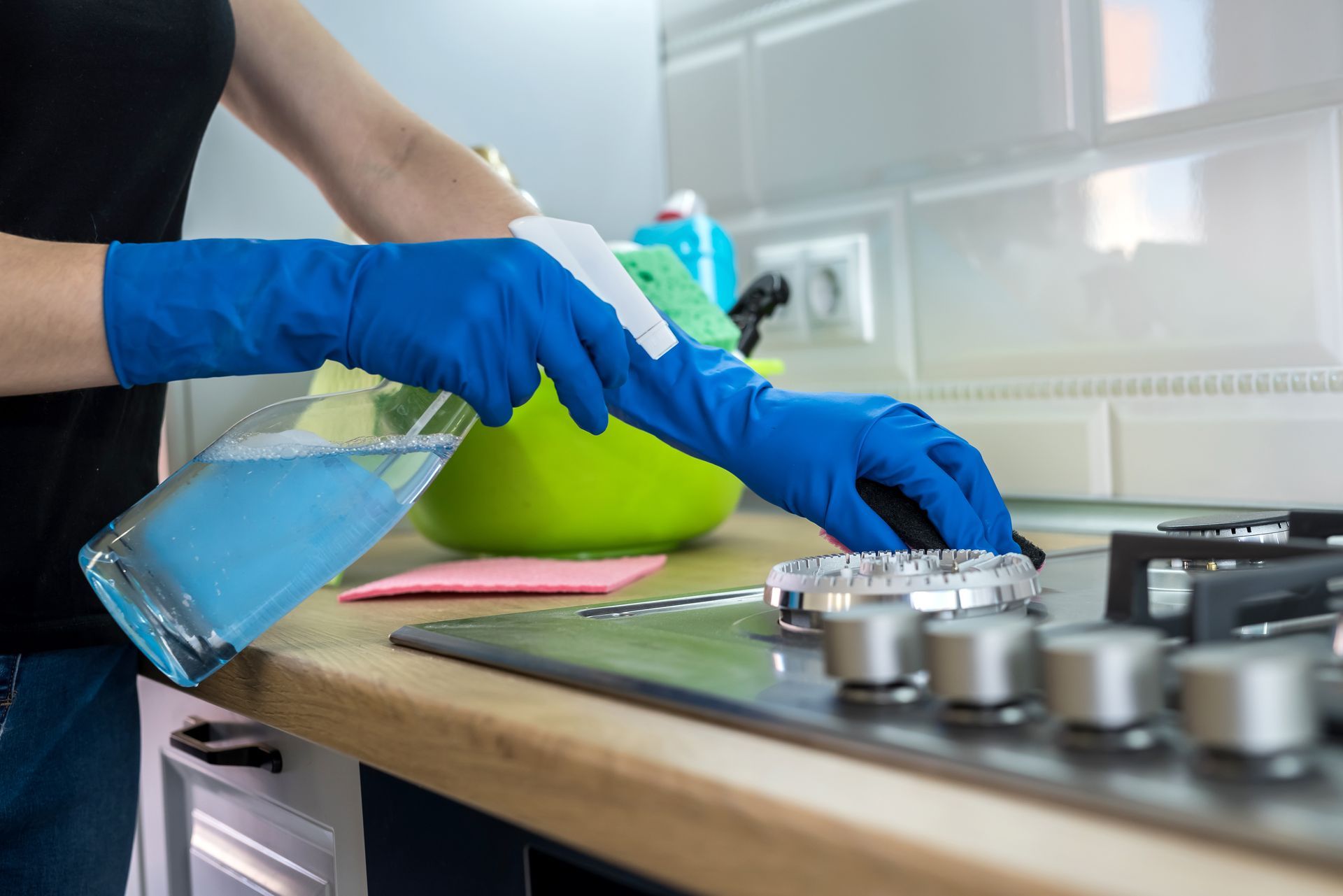 A person wearing blue rubber gloves uses a spray bottle and sponge to clean a stainless steel gas stovetop.