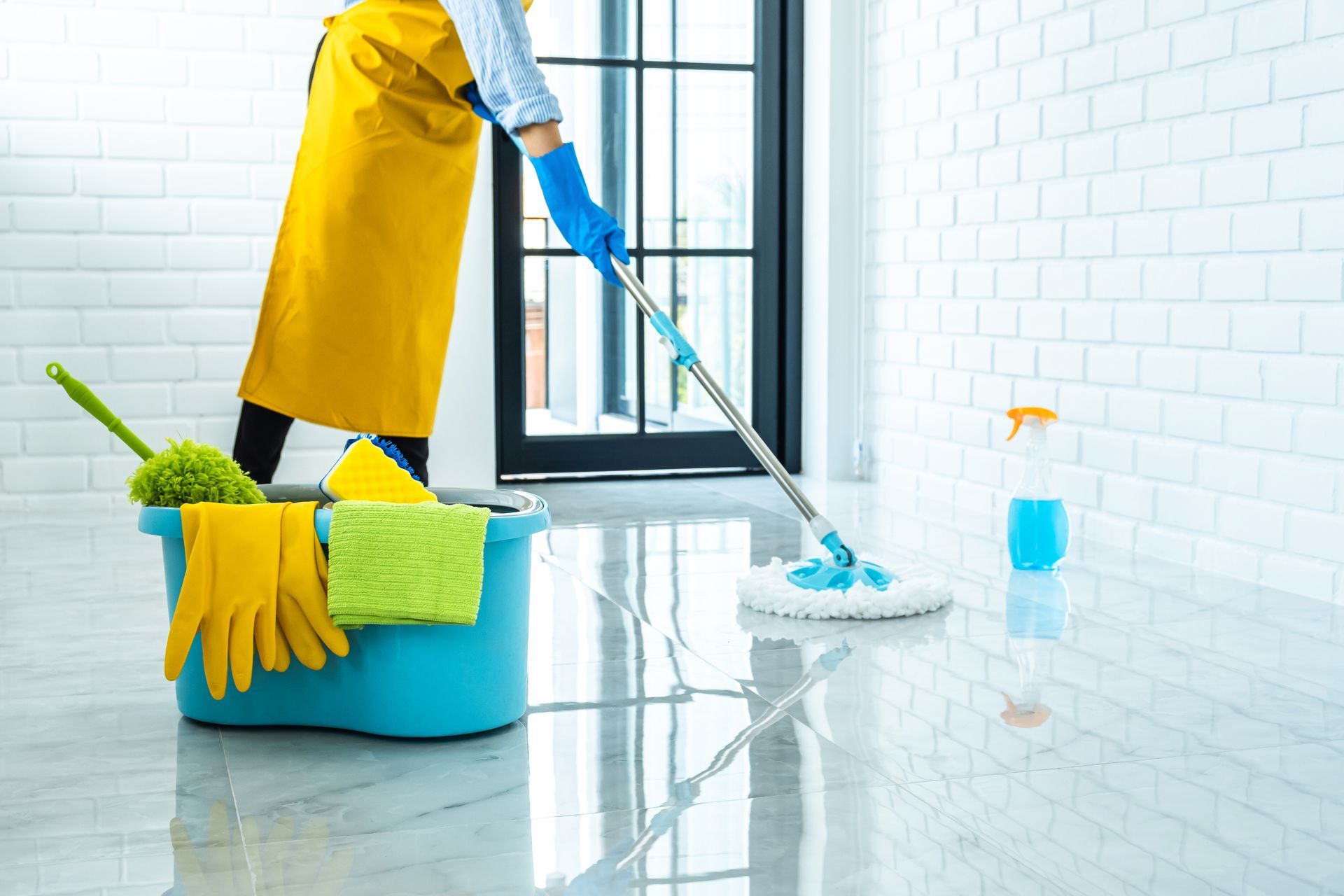 A person in a yellow apron and blue gloves mops a shiny white floor near a blue bucket filled with cleaning supplies.