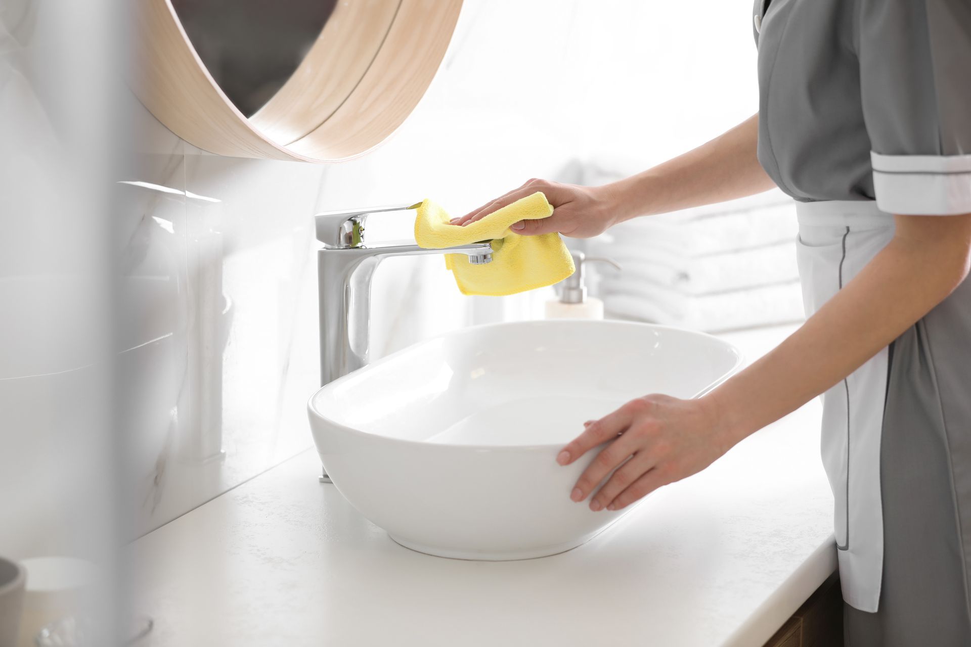 A person in a gray uniform cleans a chrome bathroom faucet with a yellow cloth above a white vessel sink.