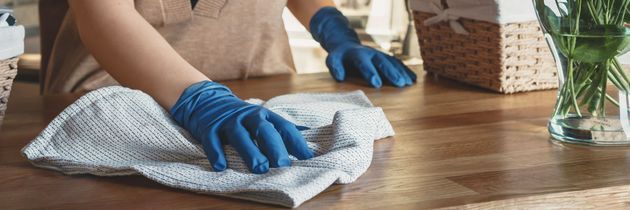 A person wearing blue gloves wipes a wooden table with a light-colored cloth.
