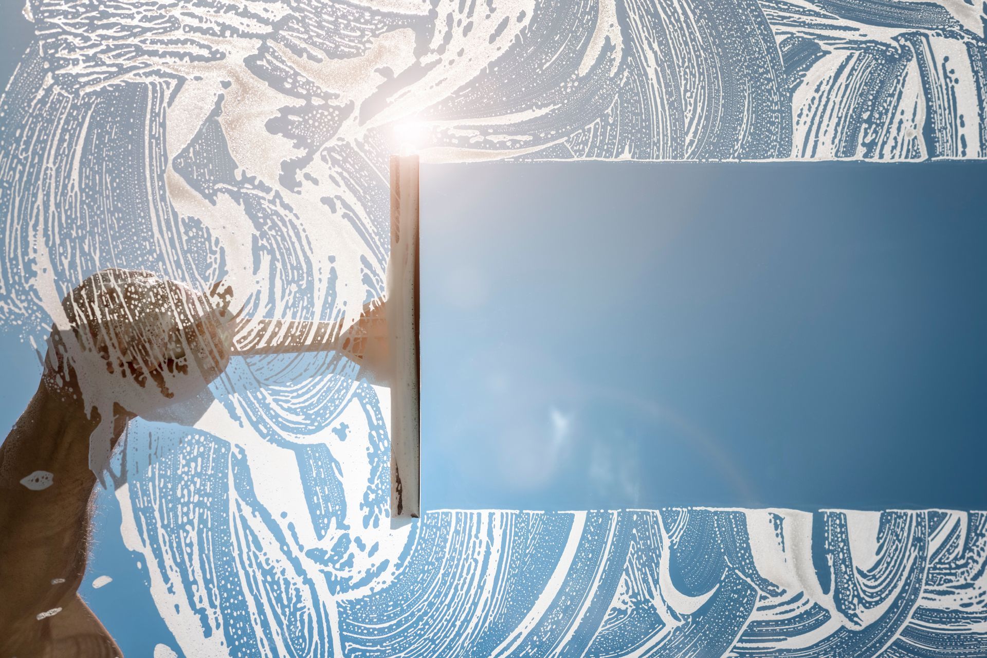 A squeegee wipes away soapy foam from a glass window, revealing a clear blue sky behind it.