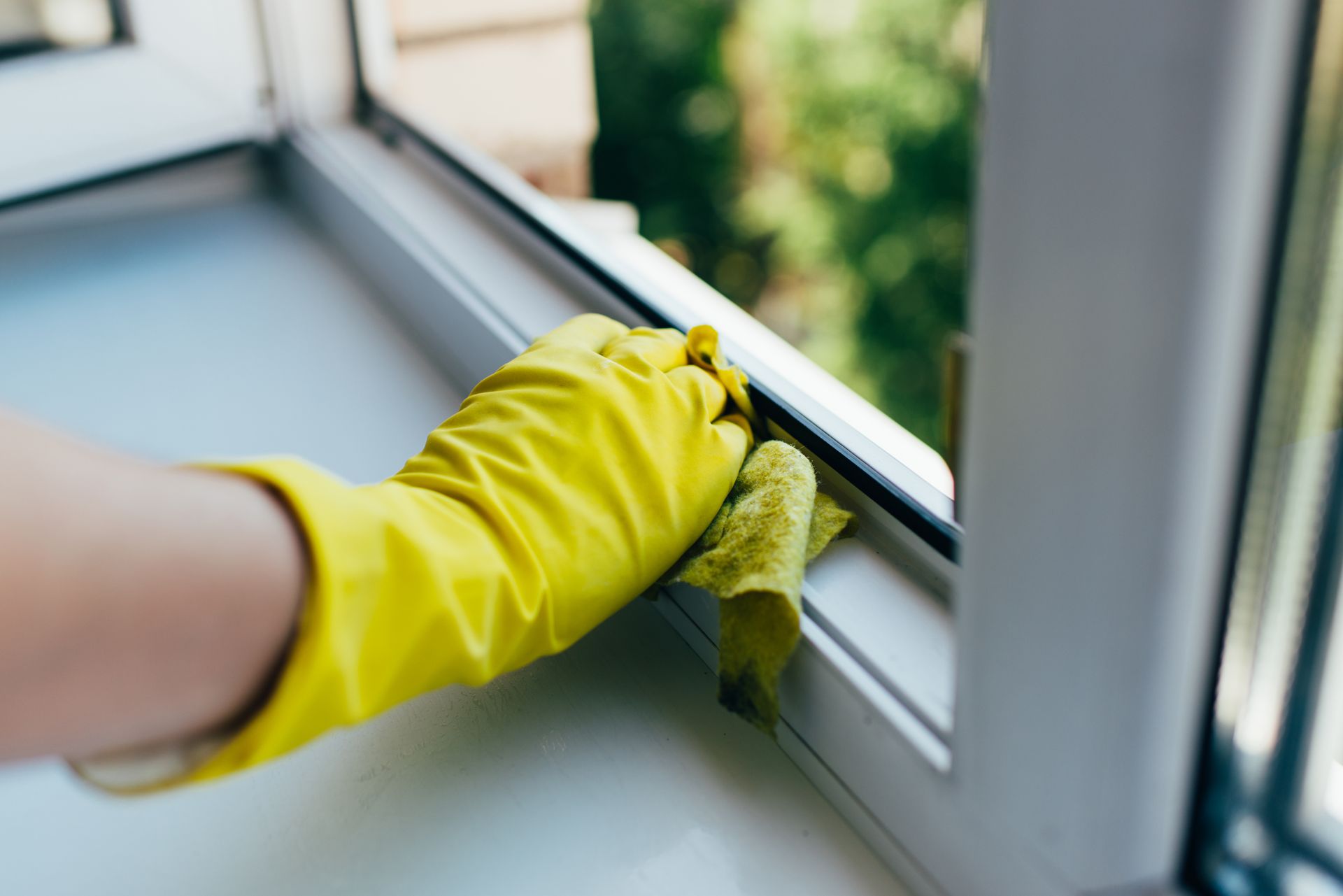A hand in a yellow rubber glove uses a cloth to wipe down a white window frame.