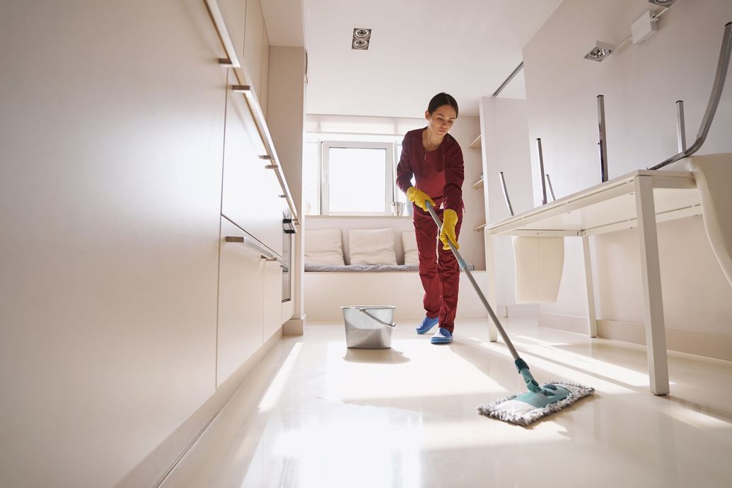 A person wearing a maroon uniform and yellow gloves mops a clean, light-colored floor in a bright, modern room.