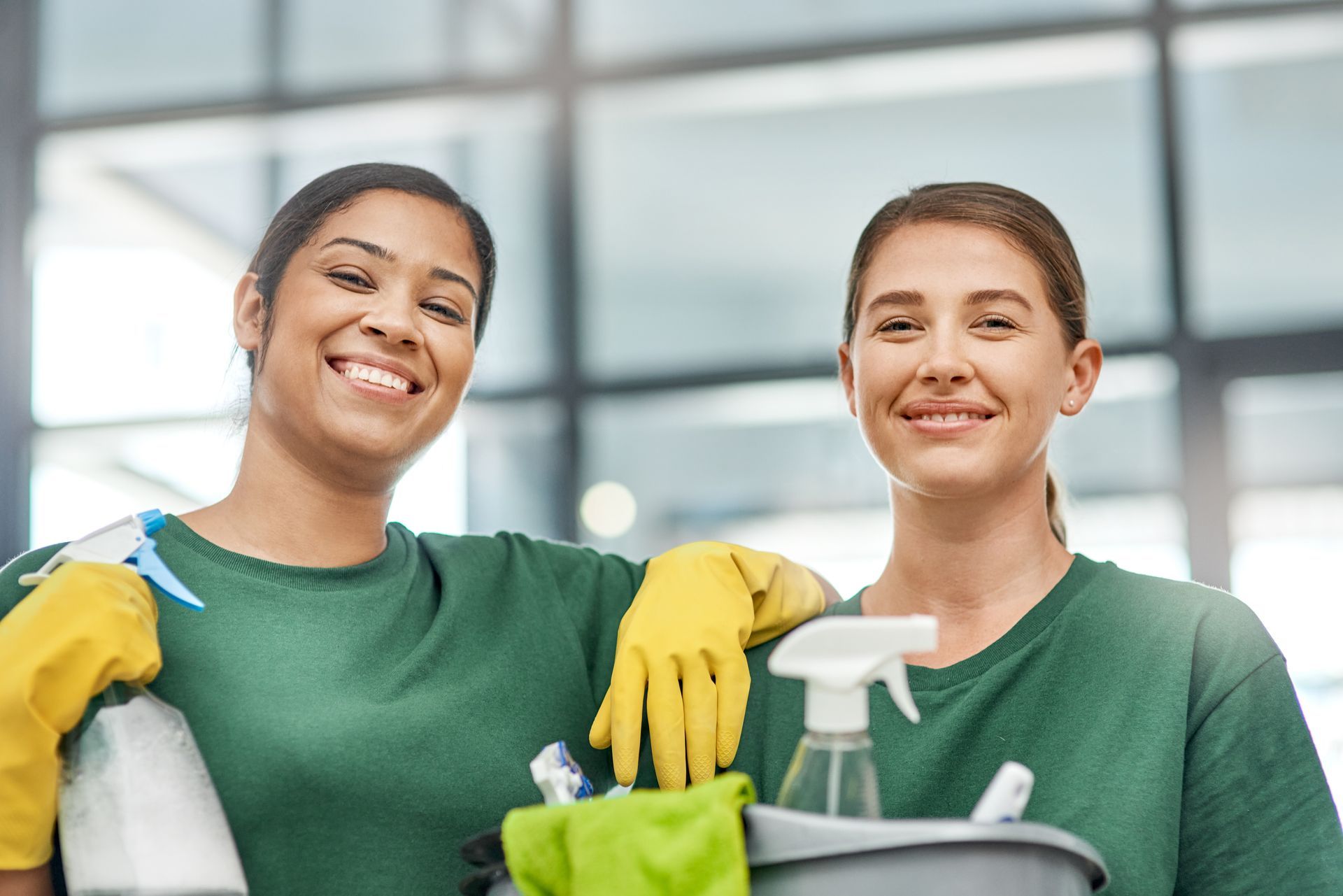 Two people wearing green shirts and yellow rubber gloves smile while holding cleaning supplies in an office setting.
