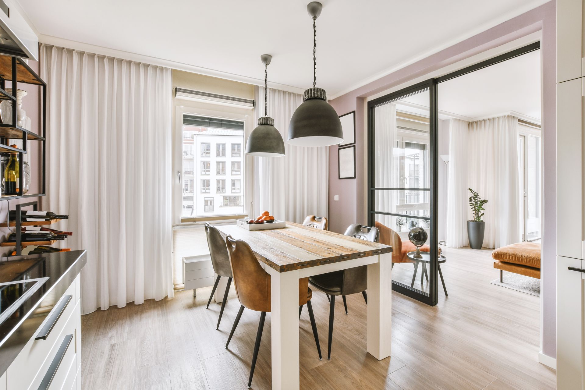 A modern dining area with a wooden table, four chairs, and pendant lights, opening into a bright adjacent living space.